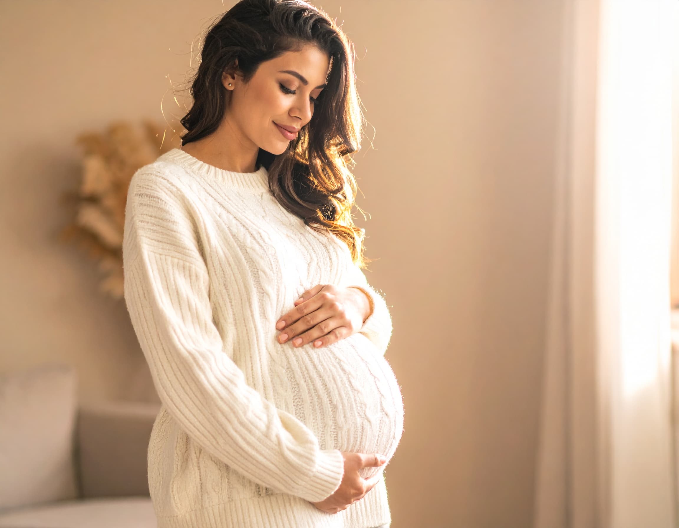 An expectant mother in a cream sweater cradling her pregnant belly in warm light.